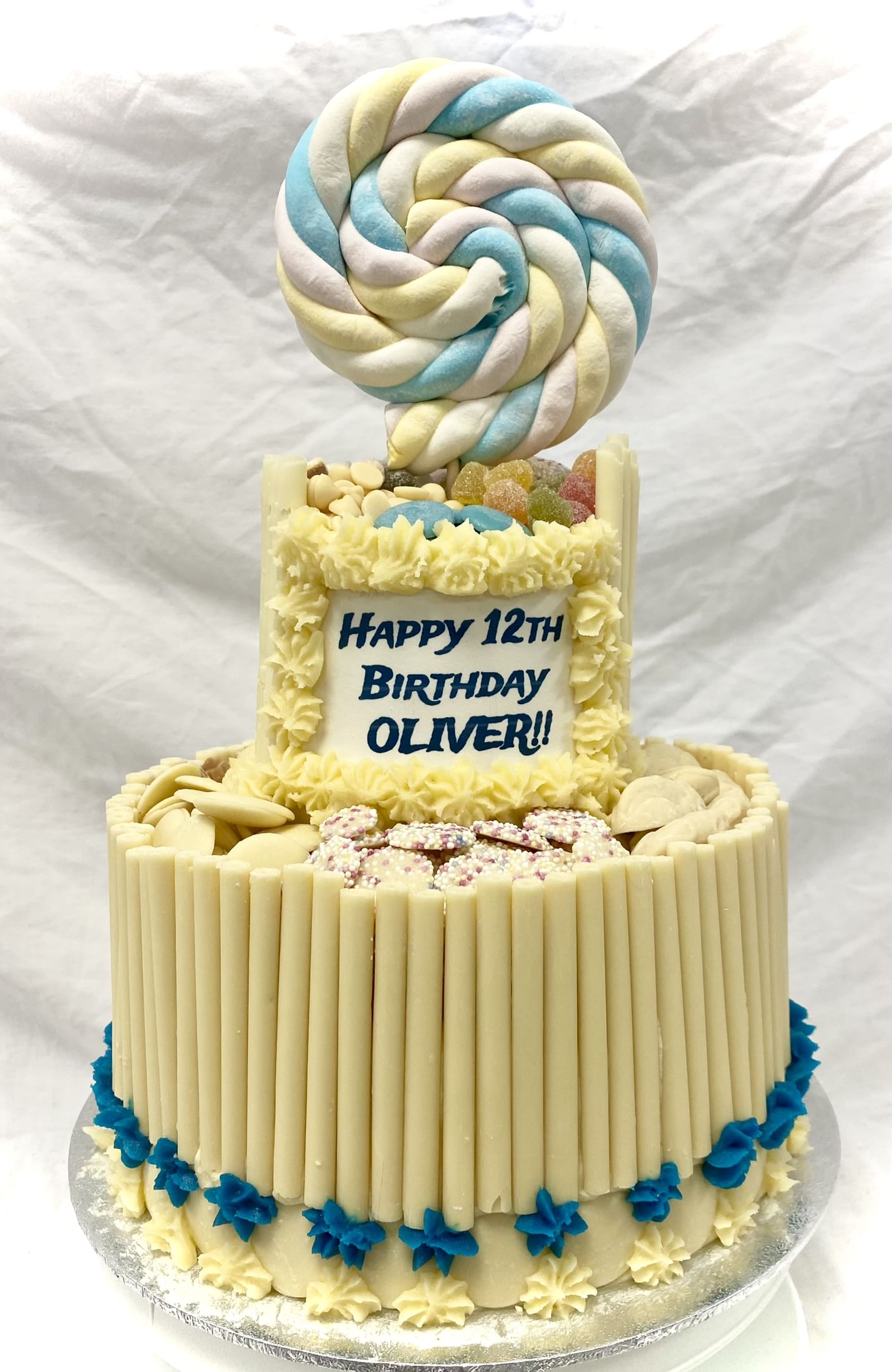 A two-tiered birthday cake decorated with white and blue icing. The bottom tier is encircled with white chocolate sticks and blue dollops of frosting, while the top tier is adorned with candies and topped with a large, swirled lollipop. This Avalanche Two-Tier Cake reads 'Happy 12th Birthday Oliver!'